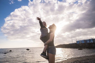 Mother carrying cute son while standing at beach against cloudy sky during sunset