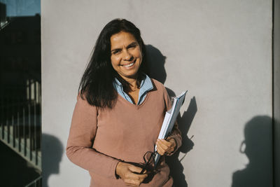 Smiling mature woman with book and file standing against gray wall at university on sunny day