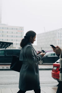 Businesswomen using mobile phones while walking on sidewalk against sky in city