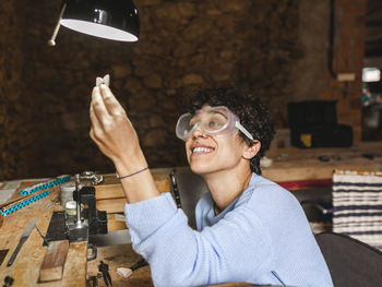 Female hispanic jeweler in glasses smiling and bringing jewelry to lamp light while sitting at workbench