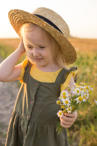 A little blonde girl in a straw hat walks in a field with a bouquet of daisies. 