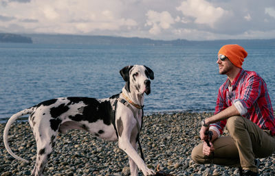 Smiling mature man with dog at beach against cloudy sky