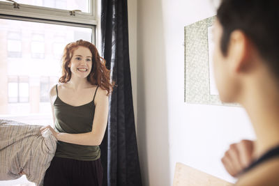 Playful lesbian with pillow looking at girlfriend while standing in bedroom