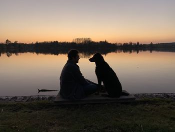 Man sitting with dog by lake against sky during sunset