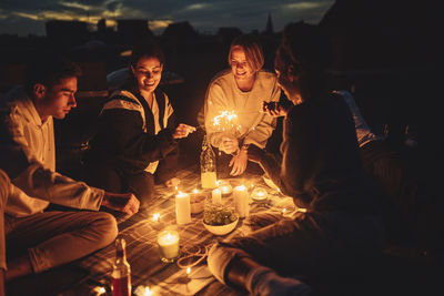 Smiling female and male friends igniting sprinkler on building terrace at night