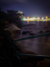 Illuminated city by sea against sky at night