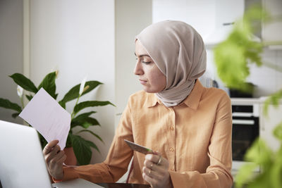 Smiling woman with hijab holding credit card while doing online shopping