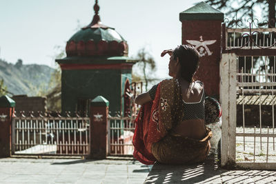 Rear view of woman sitting on footpath by fence during sunny day