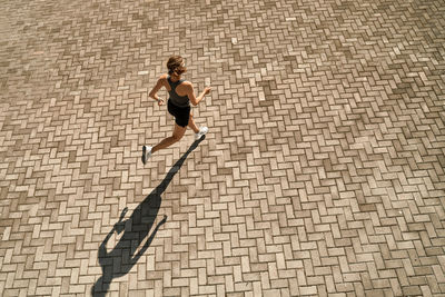 From above full body of active slim female in sportswear running fast on paved square in sunny day