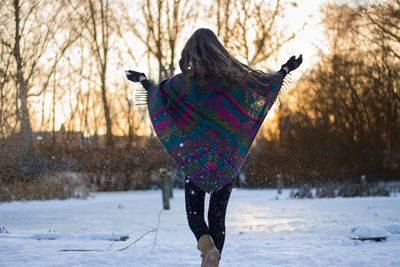 Rear view of woman wearing poncho walking against bare trees during snowfall