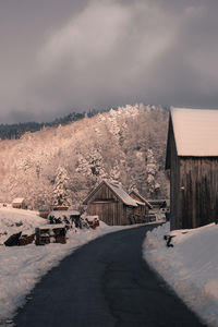 Road amidst wooden buildings against forest, mountain and sky during winter