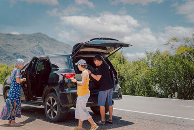 Man with female friends loading luggage in trunk of automobile on summer vacation trip.