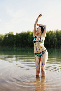 Young woman with paint on body standing in lake against sky