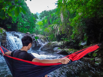 Rear view of man sitting by waterfall against trees