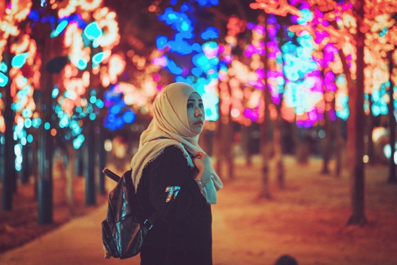 lifestyles, focus on foreground, real people, one person, night, illuminated, clothing, standing, leisure activity, tree, young adult, women, waist up, young women, looking, three quarter length, casual clothing, side view, warm clothing, outdoors, scarf