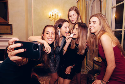 Group of positive female friends taking self portrait on smartphone while gathering at table with food during dinner in restaurant
