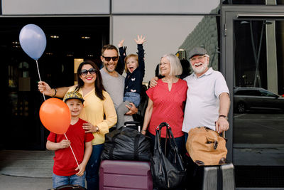 Portrait of cheerful family with luggage standing at hotel entrance during vacation