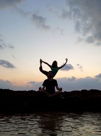 Man and woman exercising on rocks by sea against sky during sunset