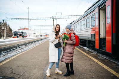 Two girls on the station platform with flowers happily looking into a smartphone