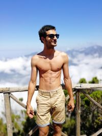 Full length of shirtless man standing by railing against sky