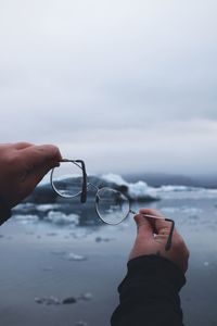 Cropped hand of man holding eyeglasses against sea