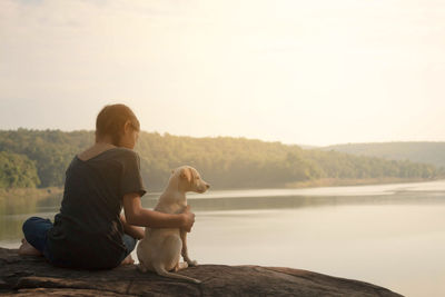 Rear view of woman with dog sitting on lake against sky