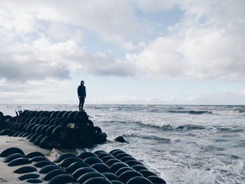 Young man standing on tire at beach against cloudy sky