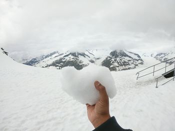 Cropped hand of person holding snow on mountain against cloudy sky