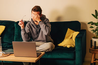 Frustrated man sitting on the green couch in his living room in front of his laptop. 