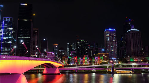 Illuminated bridge over river by buildings against sky at night