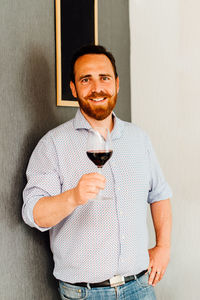 Man with moustache holding glass of red wine while standing at wall