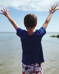 Rear view of boy with arms raised standing at beach against sky