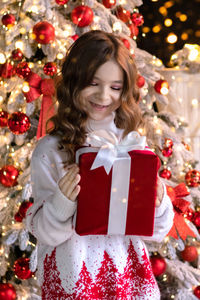 A joyful girl looks at a gift against the background of a christmas tree.