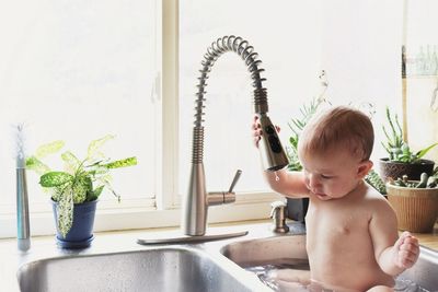 A small boy takes a bath in the kitchen sink near a sunlit window. 