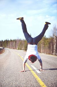 Full length rear view of man doing handstand on country road against cloudy sky