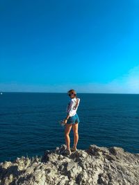 Side view of young woman standing on rock by sea against clear blue sky during sunny day