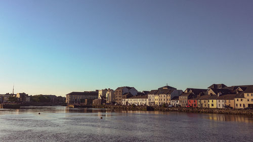 Buildings by river against clear blue sky