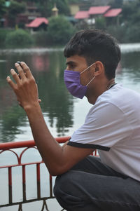Side view of a guy using mobile phone, wearing face mask sitting beside the lake