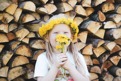 A little girl with a wreath on her head sitting near the firewood stack outdoors.