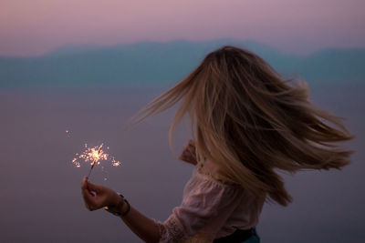 Woman holding sparkler against sky during sunset