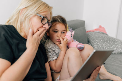 Granny and preschool girl using tablet together at home on the sofa, family togetherness time