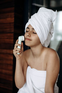 Young girl with towel on head makes facial massage with wireless electric massager