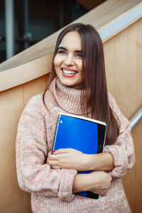 Smiling young woman holding book while standing in college