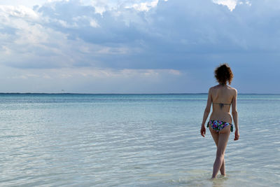 Rear view of young woman in bikini walking at beach against sky