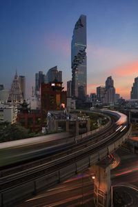 Light trails on road by buildings against sky during sunset