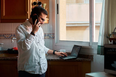 Young man calls with his smartphone and looks his laptop at kitchen