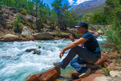 Man sitting on rock by river flowing in forest