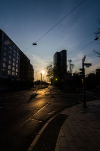 Empty road by buildings against sky during sunset