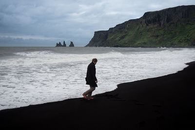 Full length of man enjoying at black beach against cloudy sky