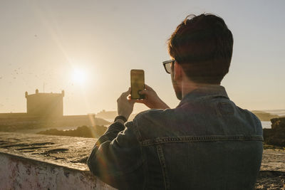 Rear view of man photographing with mobile phone against clear sky during sunset